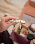 A hand holds a small tube labeled WILDFLOWER in front of an open gift box filled with assorted products, photographed in a softly lit indoor setting.