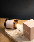 An amber glass jar with a pale pink label lies on its side on a marble slab, with white powder spilled out. A matching pale pink box sits beside it on a wooden surface against a dark background.