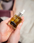 A hand holding a bottle of REVE Botanicals Body & Skin Care hair growth serum, showing the golden liquid and white dropper cap against a soft-focus background.