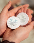 Two small round white jars of honey lavender essential oil hand and foot soaks held in a person's hands, with the front jar label clearly showing the brand and product name.