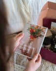 A person holds a copy of 'The Wildflower Edition' book in front of an open gift box containing various botanical and wellness items, set against a cozy background of white bedding.
