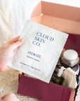 A hand holds a Cloud Skin Co. Hydrate sheet mask packet in front of an open pink gift box containing skincare items, set against a soft neutral background.