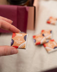 A hand holds a small square box of BBC Goodness Davidson Plum confectionery sweets, with several matching boxes blurred in the background on a light fabric surface.