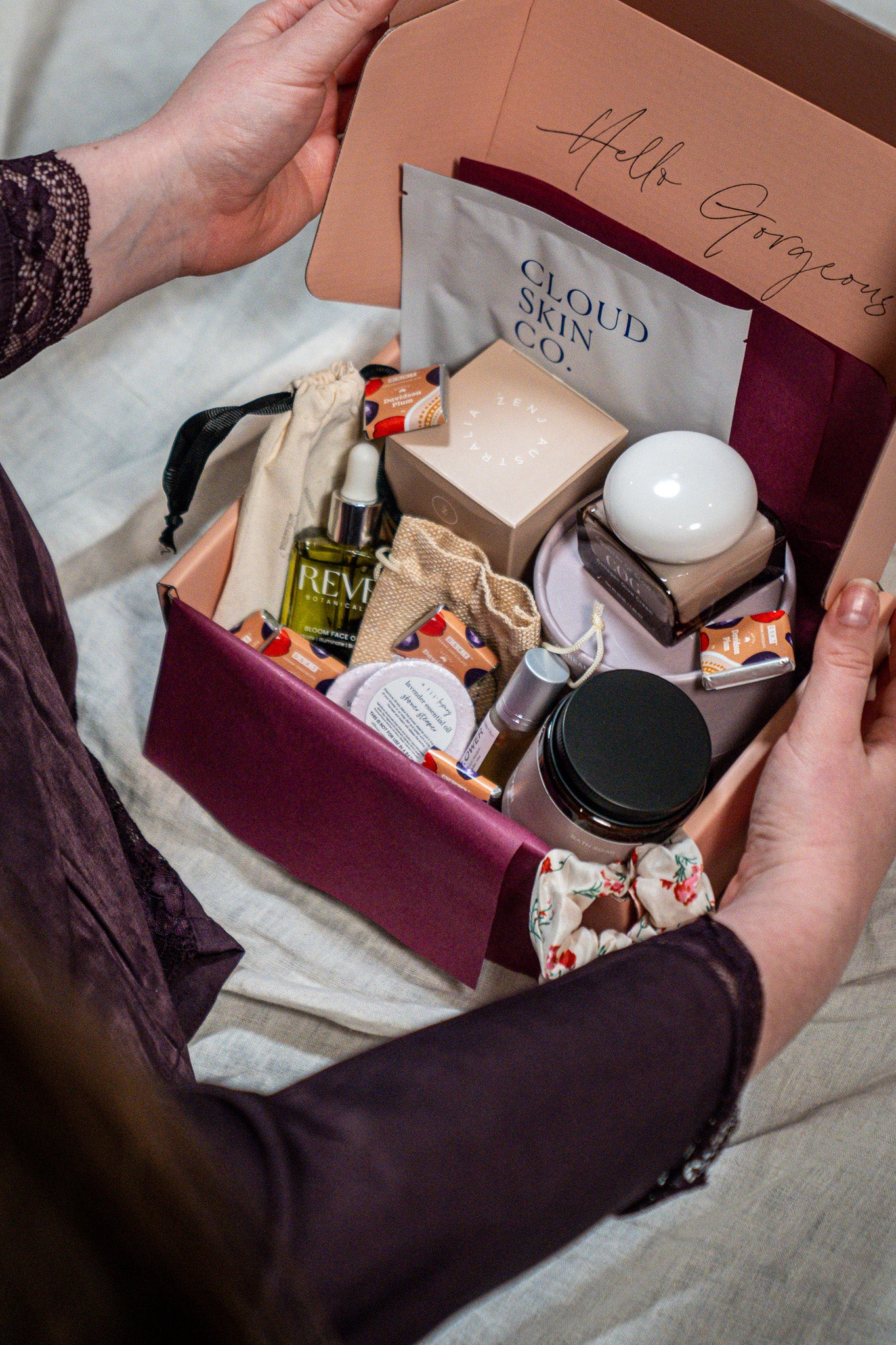 A person opens a pink gift box filled with assorted skincare and beauty products, including a Cloud Skin Co. packet, a green serum bottle, and other packaged items, set against a soft neutral fabric background.