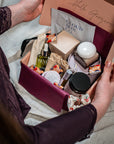 A person opens a pink gift box filled with assorted skincare and beauty products, including a Cloud Skin Co. packet, a green serum bottle, and other packaged items, set against a soft neutral fabric background.