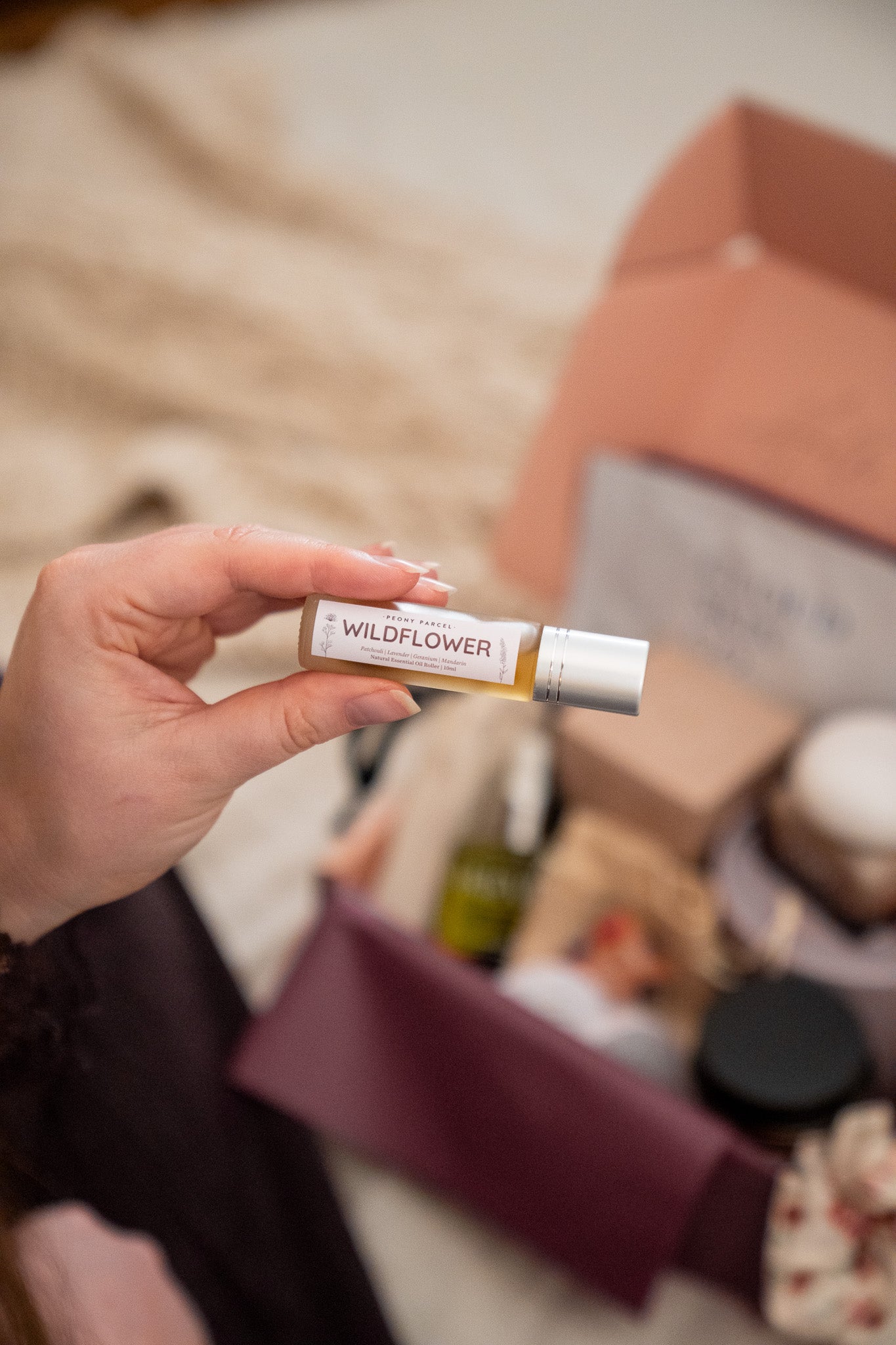 A hand holds a small tube labeled WILDFLOWER in front of an open gift box filled with assorted products, photographed in a softly lit indoor setting.