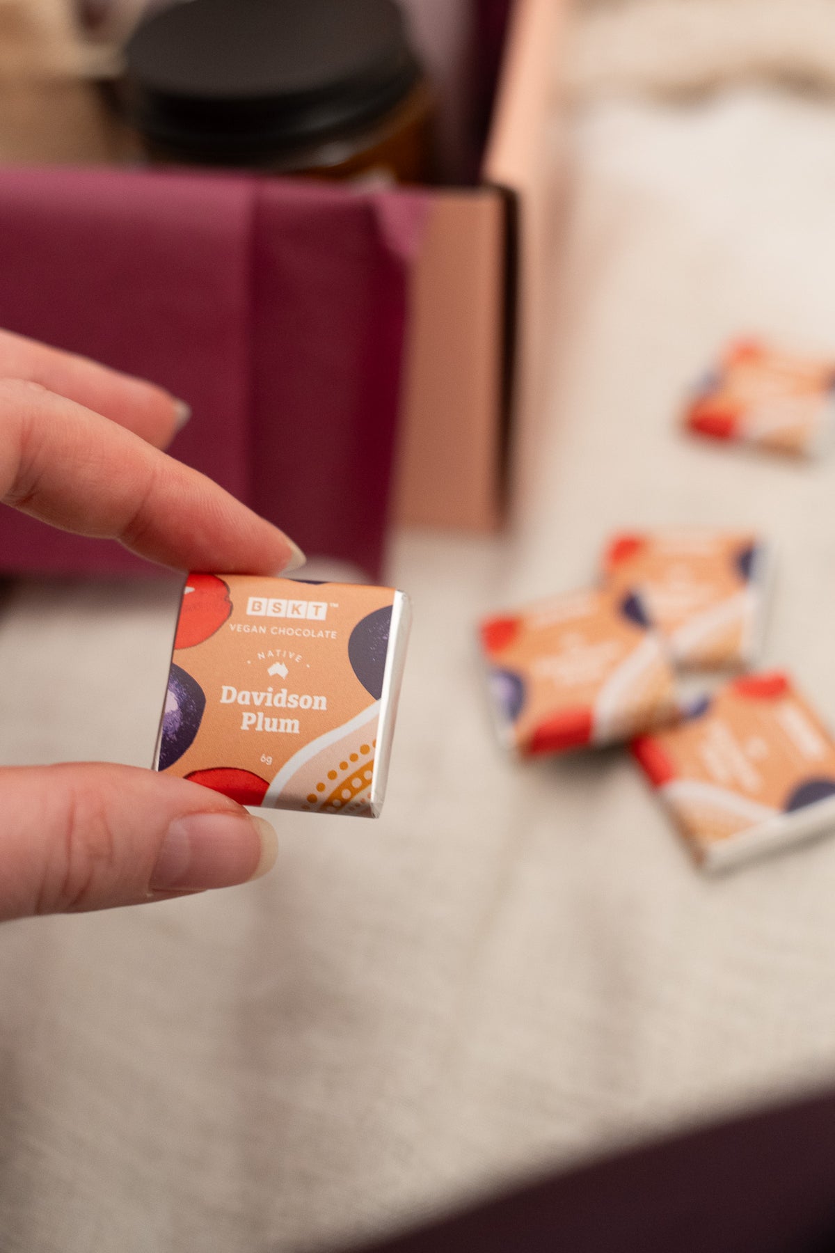 A hand holds a small square box of BBC Goodness Davidson Plum confectionery sweets, with several matching boxes blurred in the background on a light fabric surface.