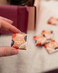 A hand holds a small square box of BBC Goodness Davidson Plum confectionery sweets, with several matching boxes blurred in the background on a light fabric surface.