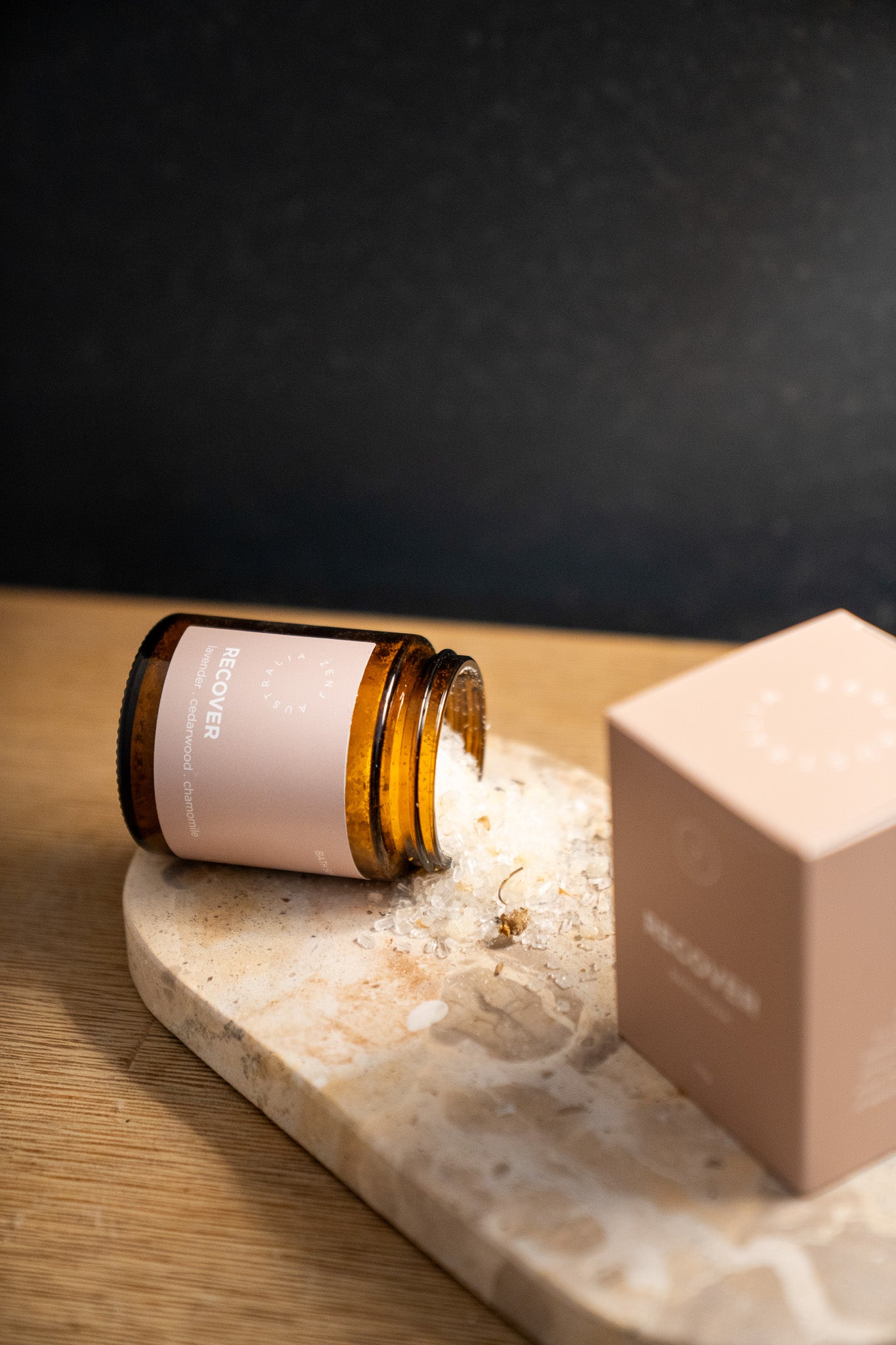 An amber glass jar with a pale pink label lies on its side on a marble slab, with white powder spilled out. A matching pale pink box sits beside it on a wooden surface against a dark background.