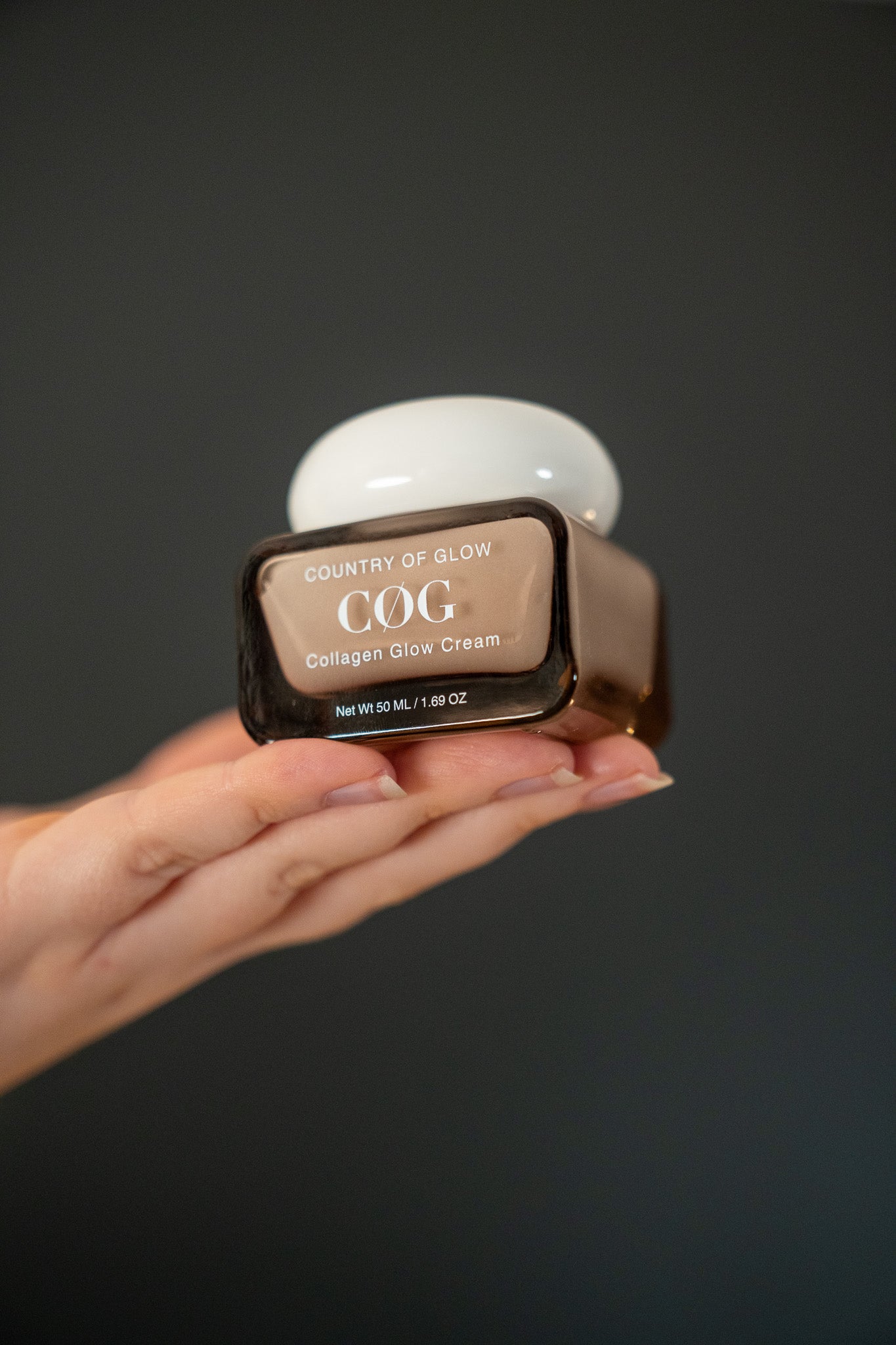 A hand holding a brown cosmetic jar of Country of Glow Collagen Glow Cream against a dark gray background.