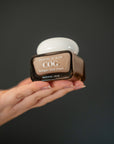 A hand holding a brown cosmetic jar of Country of Glow Collagen Glow Cream against a dark gray background.
