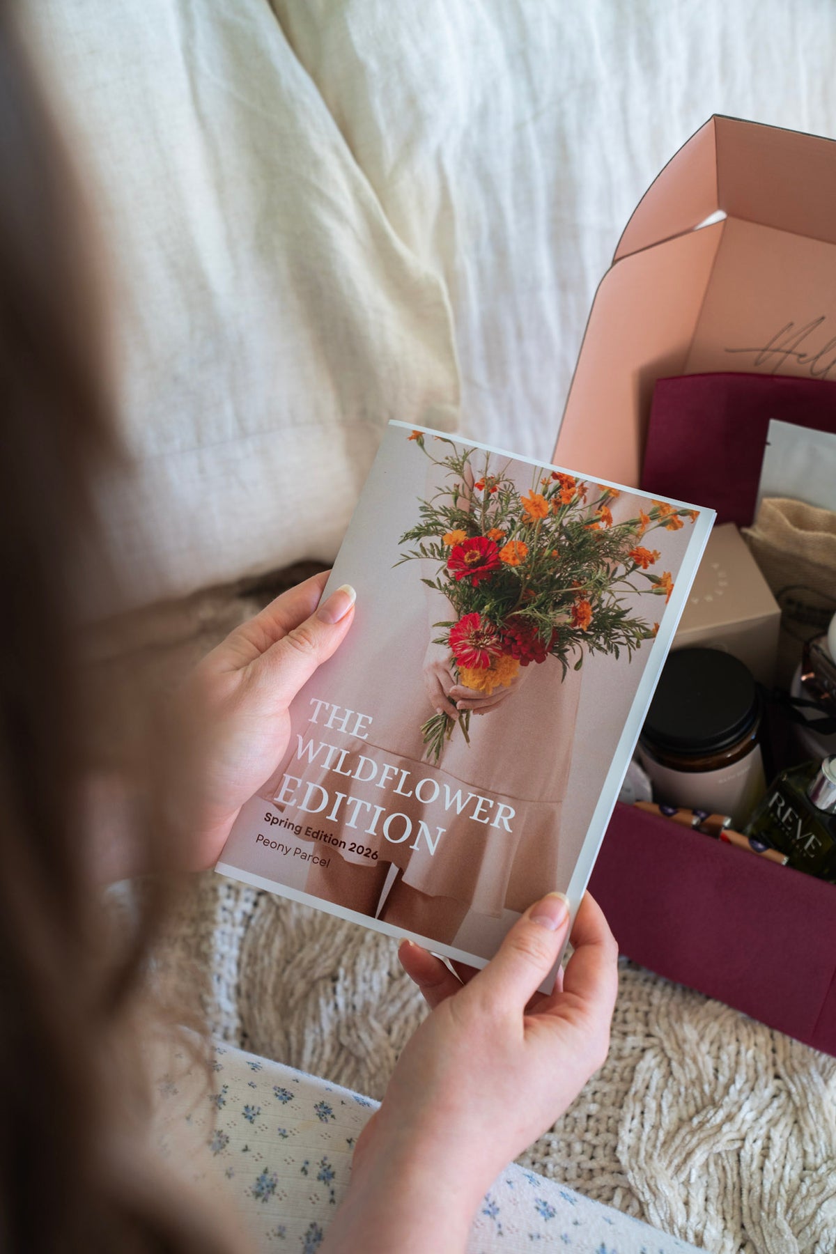 A person holds a copy of 'The Wildflower Edition' book in front of an open gift box containing various botanical and wellness items, set against a cozy background of white bedding.
