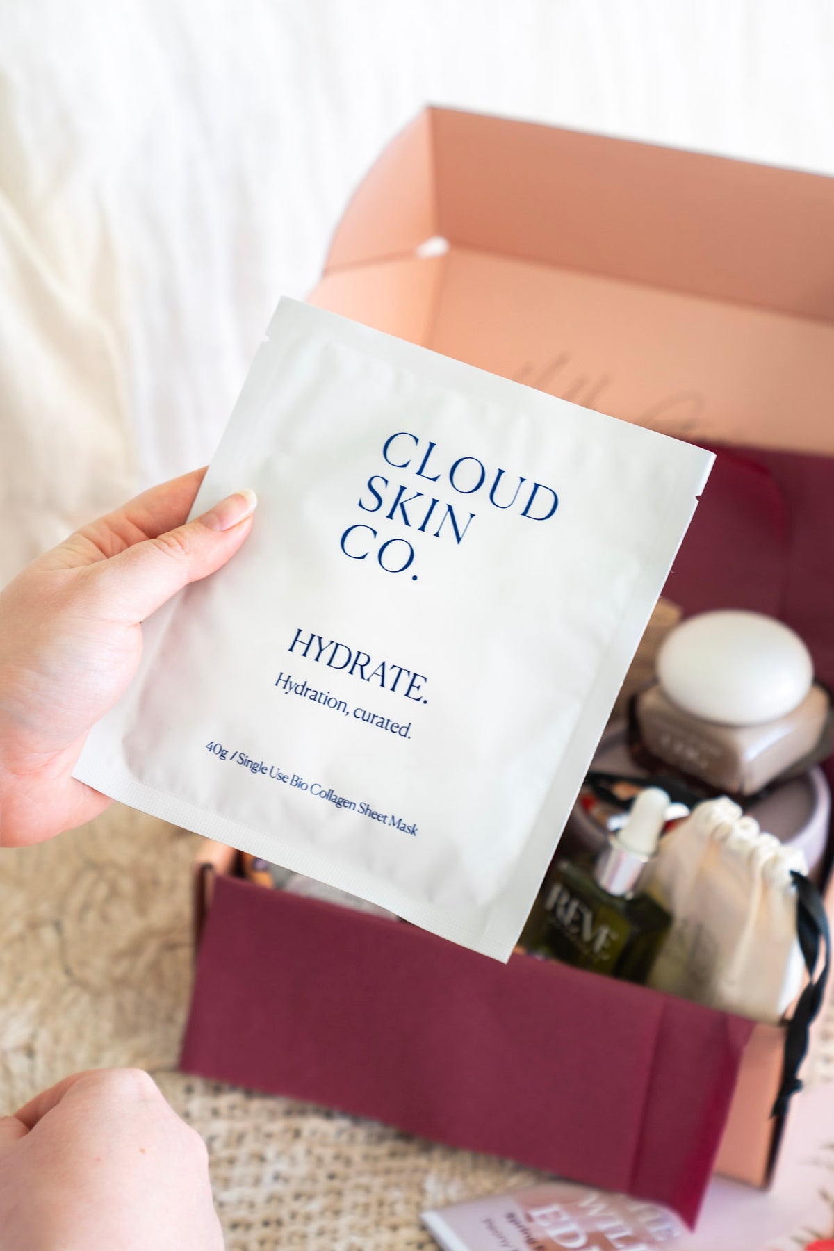 A hand holds a Cloud Skin Co. Hydrate sheet mask packet in front of an open pink gift box containing skincare items, set against a soft neutral background.