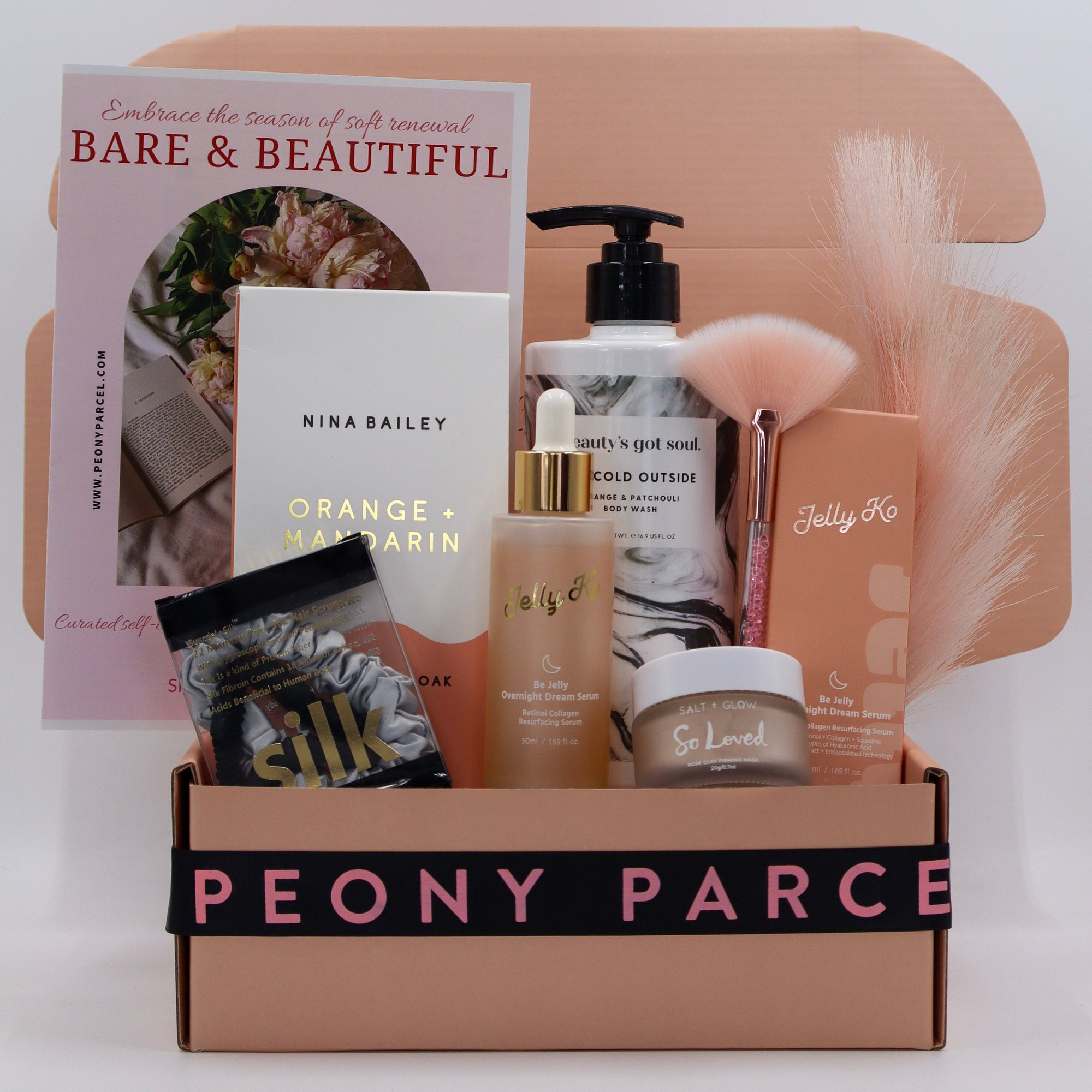 A curated beauty gift box set from Peony Parcel, featuring a collection of skincare and haircare products including a bottle of hair wax, a jar of lip balm, a soap bar, a book, and a silk pillowcase, arranged inside a beige cardboard box with a black ribbon that reads 'PEONY PARCEL'.