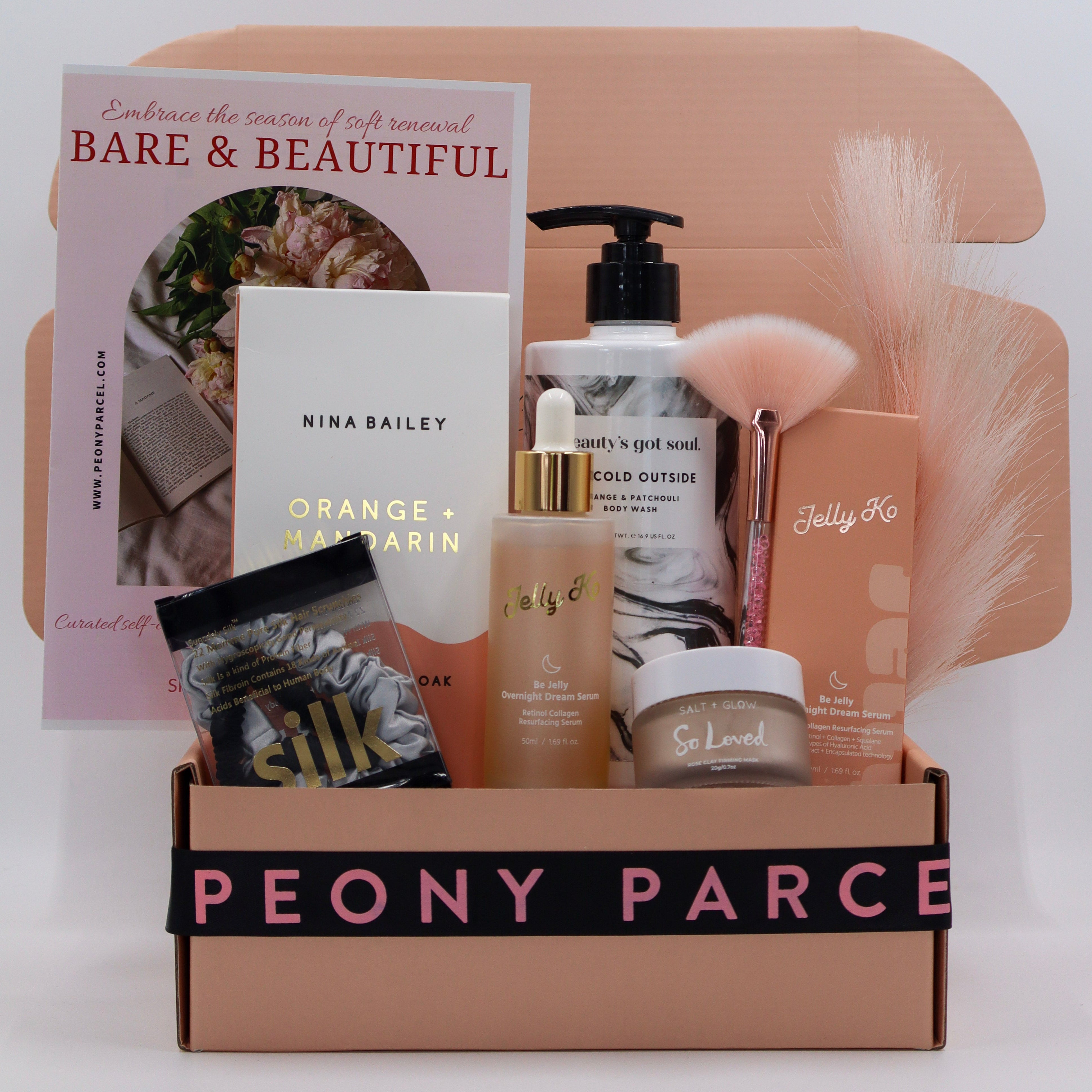 A curated beauty gift box set from Peony Parcel, featuring a collection of skincare and haircare products including a bottle of hair wax, a jar of lip balm, a soap bar, a book, and a silk pillowcase, arranged inside a beige cardboard box with a black ribbon that reads 'PEONY PARCEL'.
