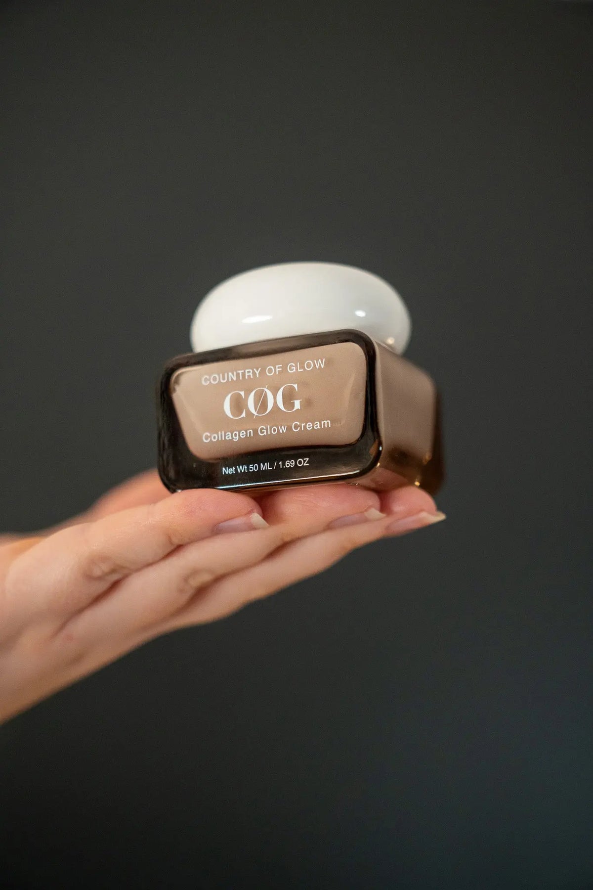 A hand holding a brown cosmetic jar of Country of Glow Collagen Glow Cream against a dark gray background.
