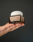 A hand holding a brown cosmetic jar of Country of Glow Collagen Glow Cream against a dark gray background.